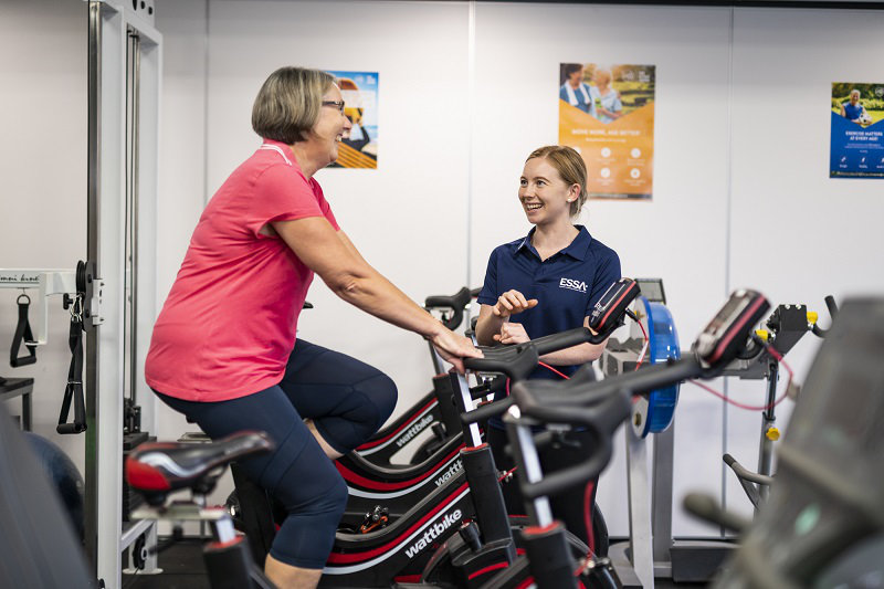 An exercise physiologist talking with an elderly woman on stationary bicycle