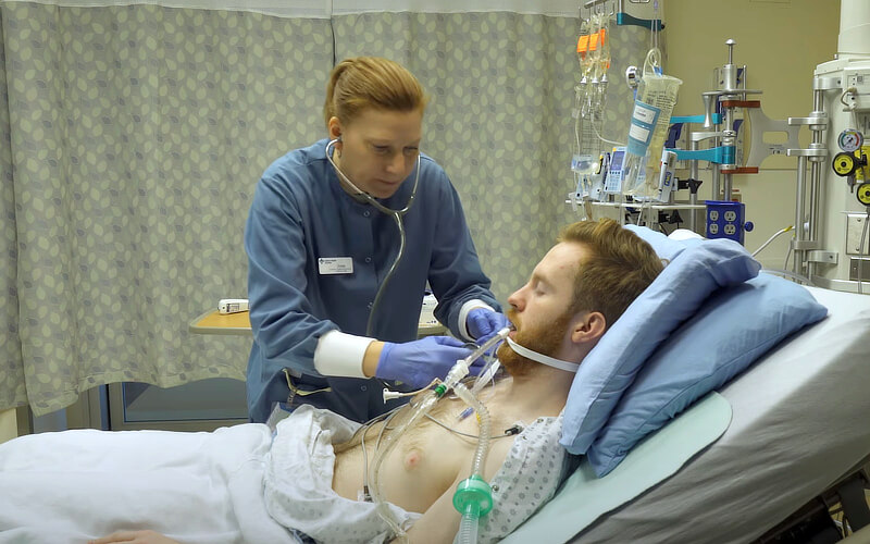 A man is lying in a hospital bed, with a tube attached to his mouth. Attended to the man is a registered nurse with a stethoscope around her neck.